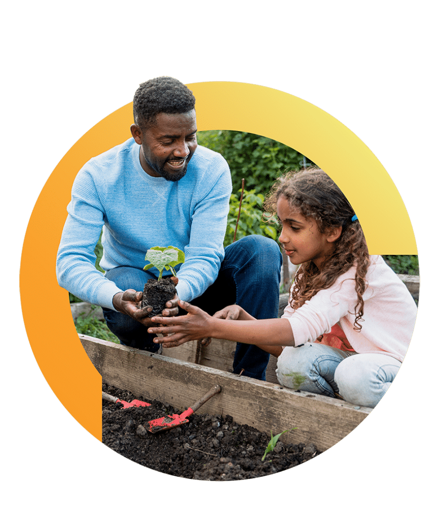 A man hands a young girl a small seedling to plant in a raised garden bed.