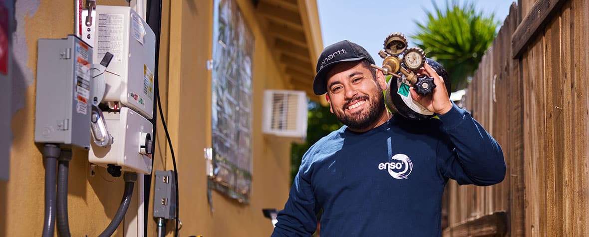 A contractor smiling outside on the side of a house and holding a piece of equipment.