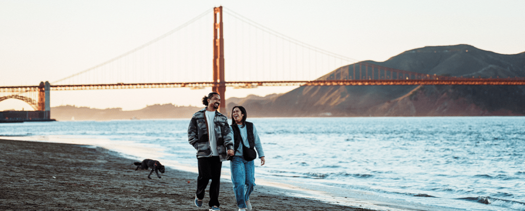 A view of the Golden Gate Bridge in San Francisco at sunset with a couple and a dog walking on the beach in the foreground.