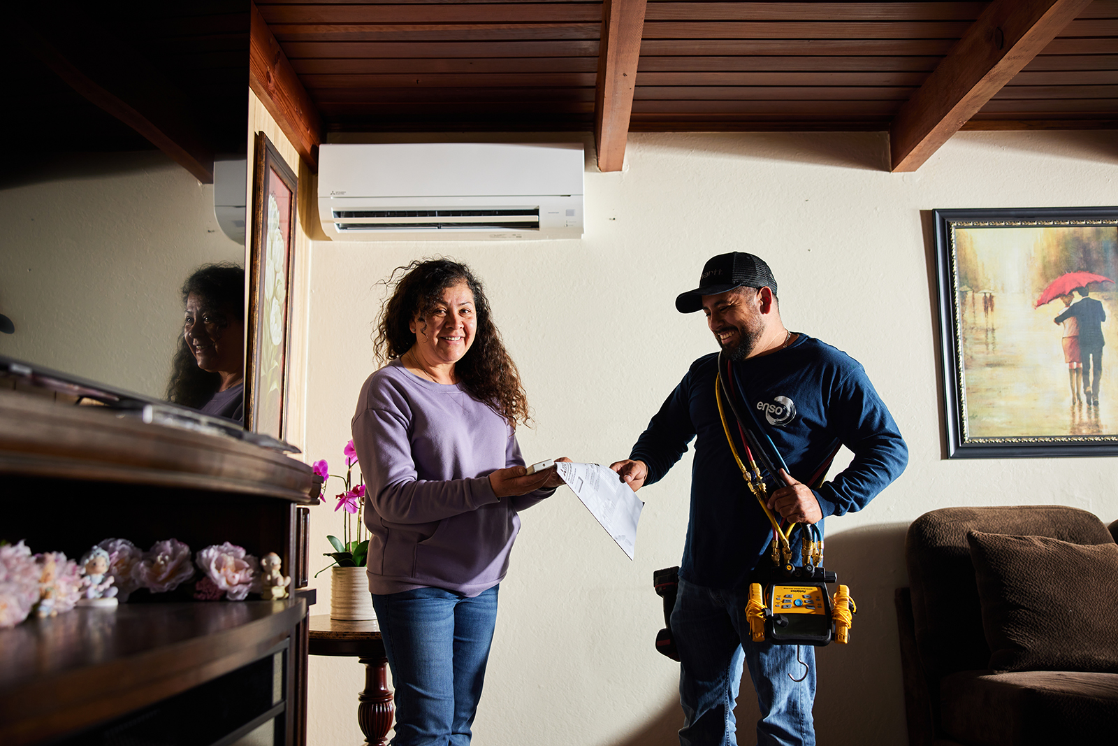 A woman smiles while handing paperwork to a technician carrying tools. They're indoors by an air conditioning unit and framed artwork. Warm, friendly atmosphere.