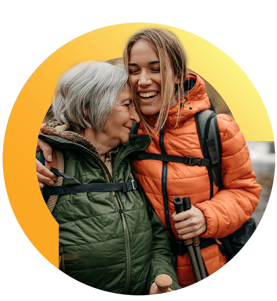 An older woman and younger woman embrace and smile together while dressed for hiking outdoors.