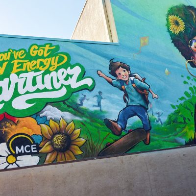 A colorful large-scale mural on an exterior building wall reads "You've Got Good Energy Martinez" in bold yellow and white lettering on a green background. The mural features a joyful young boy skateboarding through a green hillside landscape with kites flying, vibrant sunflowers and red flowers in the foreground, the MCE logo, and a partial view of a joyful woman with an afro, sunglasses, and gold hoop earrings to the right of the boy.