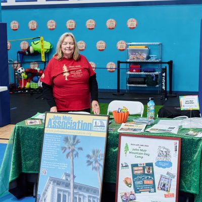 A woman with shoulder-length blonde hair wearing a red John Muir Day Camp t-shirt stands behind a table draped in green cloth at an indoor community fair. The table displays two framed posters for the John Muir Association and John Muir Mountain Day Camp, along with brochures, an orange pumpkin-shaped candy dish, and a water bottle. A Wall of Champions banner with blue jersey graphics is visible behind her.
