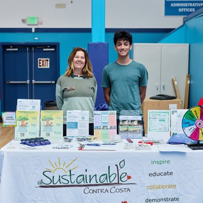 A woman with shoulder-length auburn hair wearing a green pullover and a teenage boy in a teal t-shirt stand together behind a white-tableclothed table. The table is filled with brochures, seed packets, pens, and informational materials. A colorful prize wheel sits on the right side of the table. The tablecloth reads "Sustainable Contra Costa" with the tagline "inspire, educate, collaborate, demonstrate." A "Gold Medalists" honor board is visible on the wall behind them.