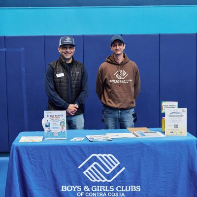 Two men wearing baseball caps stand behind a blue-draped table inside a gymnasium. The man on the left wears a black puffer vest and a Boys & Girls Clubs cap; the man on the right wears a brown Boys & Girls Clubs of Contra Costa hoodie. The table has an "After-School Program" flyer, a "Planting Day" event sign, and various informational materials. Blue gymnastics wall padding is visible in the background.