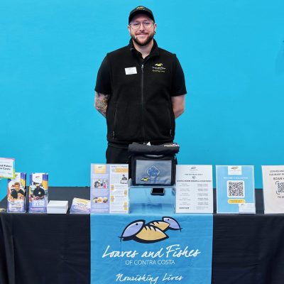 A bearded man with glasses and a black cap wearing a black Loaves and Fishes vest stands behind an info table inside a gymnasium. The table is covered with a black cloth and a light blue Loaves and Fishes banner reading "Nourishing Lives." Displayed materials include a lunch pail bag, dining room hours signage, a "Become a Volunteer" flyer, and program brochures. A teal wall is visible behind him.