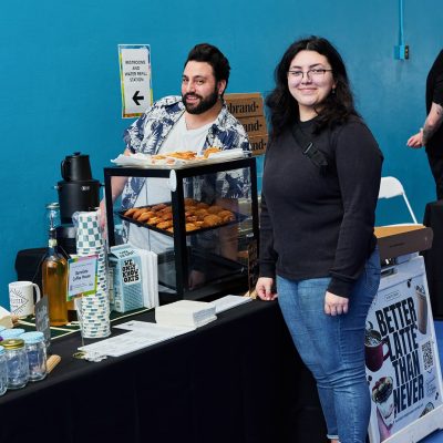 A man in a floral shirt stands behind a black-draped table set up as a coffee and pastry station, with croissants displayed on a tiered rack, mason jars, cups, and coffee equipment. A young woman with dark curly hair and glasses in a black top stands to his right smiling at the camera. A sign on the table reads "Barrelista Coffee House" and a sandwich board advertises "Better Latte Than Never."
