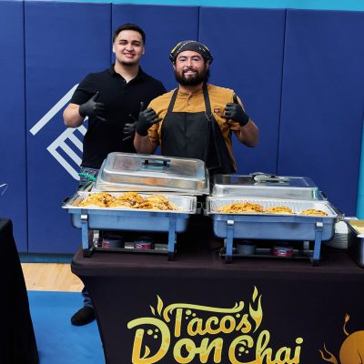 Two men stand behind a black-draped table with chafing dishes filled with tacos at an indoor event. The man on the left wears a black polo shirt and black gloves and gives a thumbs up. The man on the right wears a mustard-yellow shirt, a black apron, a black bandana, and black gloves, also giving a thumbs up. The table skirt reads "Tacos Don Chai – Catch us in action on Instagram @TacoDonChai."