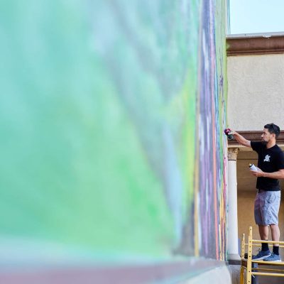 An artist in a black t-shirt and gray shorts stands on a yellow rolling scaffold platform, spray painting the upper section of a large mural on a building exterior. He holds a red spray can and reaches toward the wall. Additional spray paint cans are visible on the scaffold below him. The mural behind him shows vibrant greens and rainbow-hued colors in progress. Classical columns of a building are visible to the right.
