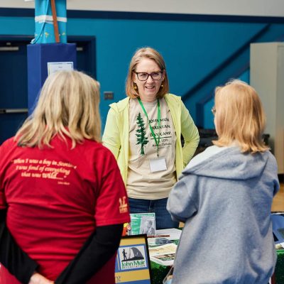 A woman with shoulder-length reddish-blonde hair wearing glasses, a lime green jacket, and a John Muir Mountain Day Camp t-shirt with a green lanyard stands behind a resource table, smiling warmly while speaking with two attendees facing away from the camera. A Boys & Girls Club administrative office sign and a "Go Team" banner are visible in the background.