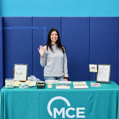 A smiling woman with long dark hair wearing a gray MCE zip-up sweatshirt waves at the camera while standing behind a teal-draped table. The table displays an MCE framed poster reading "You Have Good Energy, Martinez," pamphlets, branded merchandise, a candy dish, and a "Notice of Filming" sign. The tablecloth reads "MCE mceCleanEnergy.org." Blue gymnastics padding covers the gymnasium wall behind her.