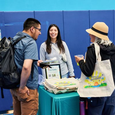 A smiling woman with long dark hair in a gray MCE sweatshirt stands behind a teal MCE table, handing materials to a young man with a small black-and-white dog peeking out of a backpack. A woman in a straw hat holding an MCE tote bag reading "You've Got Good Energy!" stands to the right. MCE informational materials and framed signs are visible on the table.