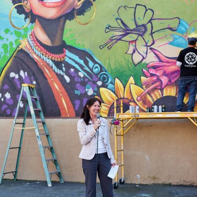 A woman with dark shoulder-length hair wearing a light blazer and gray slacks stands in front of the large mural-in-progress, holding a microphone and smiling. Behind her, the partially completed mural shows a joyful Black woman's face in vibrant color. An artist in a black Illuminaries t-shirt can be seen actively painting on the wall in the upper right corner. A green ladder and yellow rolling scaffold are visible on either side.
