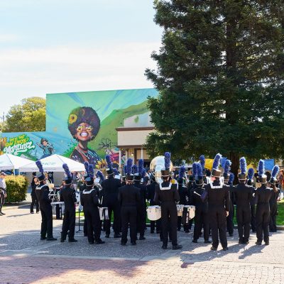 Marching band in black uniforms with blue plumes performs in a sunny plaza. The Martinez mural and large tree provide a colorful, lively backdrop.