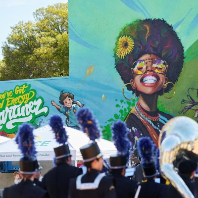 A marching band in dark uniforms with blue plume hats is seen from behind, facing the large nearly-completed "You've Got Good Energy Martinez" mural. The mural displays a joyful Black woman with an afro, sunflowers, a skateboarding boy, and kites against a sky-blue and green background. An MCE canopy tent is visible beneath the mural, and leafy trees frame the upper left.