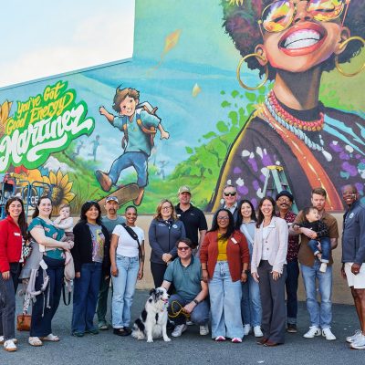 A group of approximately 17 people, including adults and two babies, pose together in front of the completed "You've Got Good Energy Martinez" mural outdoors on a sunny day. The colorful mural spans the full background, featuring a joyful Black woman with sunflowers, a skateboarding boy, and vibrant flowers. A border collie sits in front of the group. A blue aerial lift is visible to the left.
