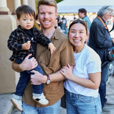 A young family of three stands outdoors in front of a blurred background showing an event with canopy tents and trees. A tall man with red hair and a brown button-up shirt holds a toddler in a plaid jacket on his hip. A smiling woman of Asian descent in a white t-shirt and jeans stands beside them with her arm around the man. The toddler holds a small piece of food.