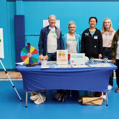 Five volunteers — four older adults and one younger woman — stand behind a blue-draped table at an indoor community fair. A colorful prize wheel and pencil cup are on the table, along with flyers and materials. To the left stands an easel-mounted sign for "350 Contra Costa Action" listing priorities including fossil fuel resistance, legislative action, and youth climate action. Several people wear 350 Contra Costa Action t-shirts.