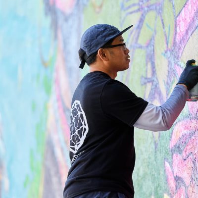 Close-up of a male muralist wearing a black Illuminaries t-shirt, a backwards cap, and glasses, concentrating as he spray paints details on the large mural wall. He wears black gloves and holds a green spray can in his right hand. The partially completed mural around him shows pink, lavender, and teal floral forms.