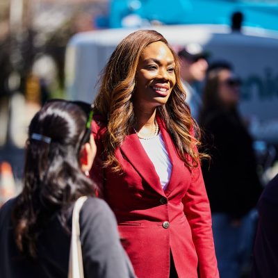 A woman in a red blazer with long auburn-highlighted hair and a pearl necklace smiles while engaged in conversation with two attendees outdoors. She is shown in sharp focus while the background features blurred figures and a blue vehicle at what appears to be an outdoor public event on a sunny day.