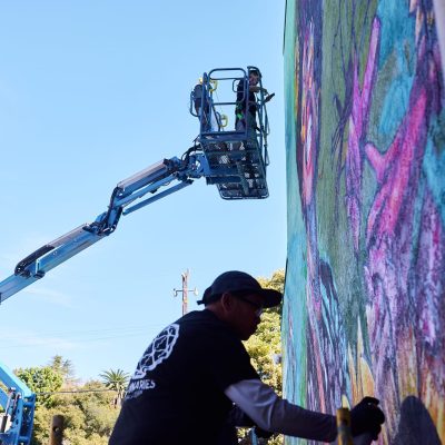 Two muralists from the Illuminaries collective work simultaneously on a tall exterior building wall covered in a vibrant mural-in-progress. One artist is elevated in a blue aerial lift cherry picker, painting the upper section against a clear blue sky. The other artist, wearing a black Illuminaries t-shirt and cap, works at ground level on the lower section. The mural in progress shows pink, teal, and green forms.