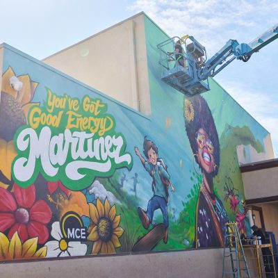 A wide-angle view of the nearly completed "You've Got Good Energy Martinez" mural on the exterior of a building, showing the full composition with colorful sunflowers, a skateboarding boy, the MCE logo, a joyful Black woman with an afro and sunglasses, and lush greenery. A blue aerial lift with an artist in the bucket is visible in the upper right still finishing the top of the mural. A yellow ladder and scaffolding with small figures stand at the base. The sky is partly cloudy.